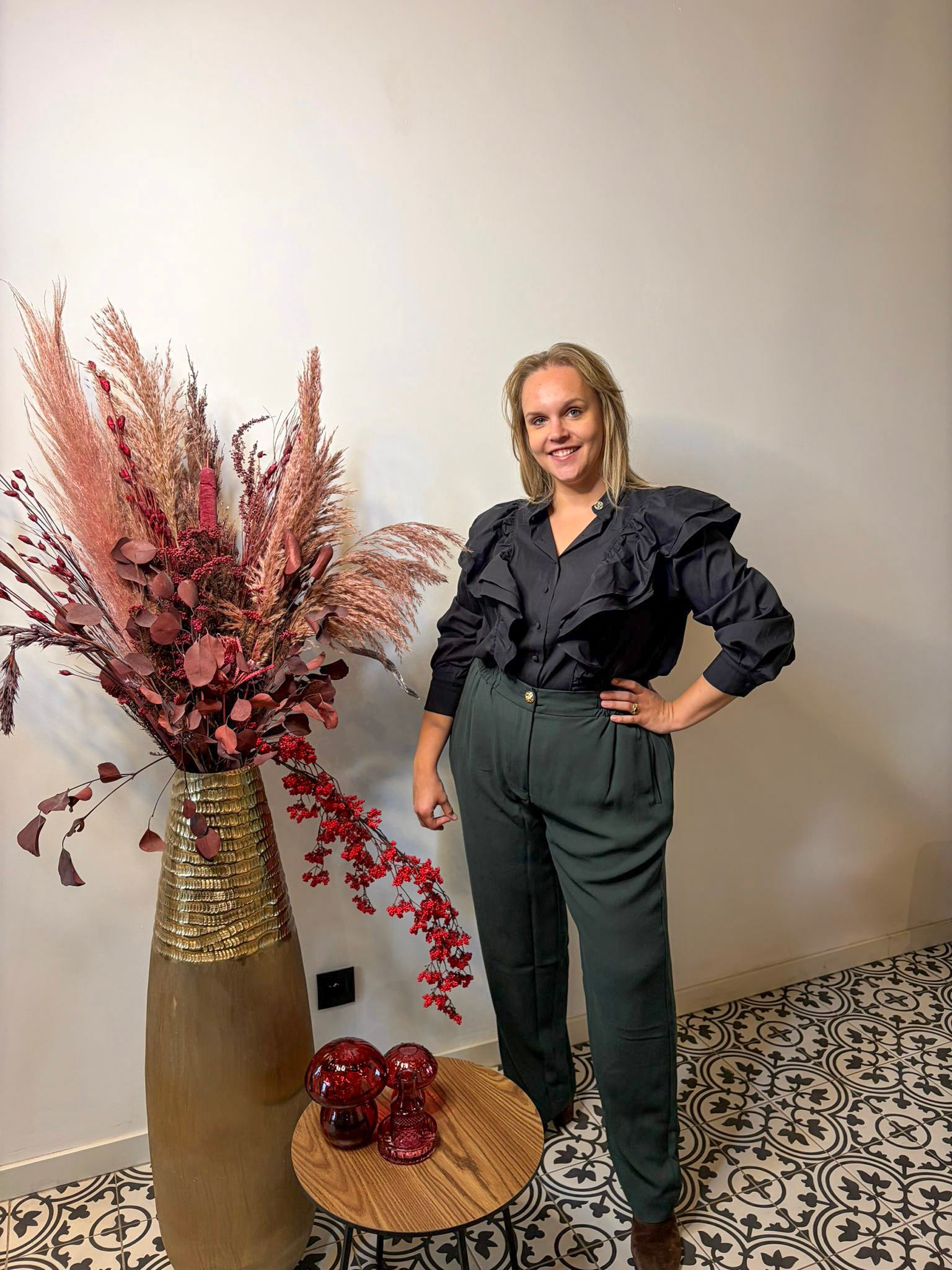 Een vrouw in een Zwart hemd - Mat Fashion met franjé-effect en een donkergroene broek met hoge taille staat glimlachend bij een houten tafeltje en een grote vaas met gedroogde bloemen op tegels met een patroon - een perfecte feestoutfit.