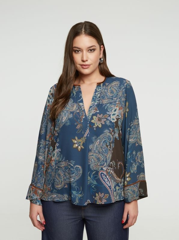 Een vrouw met lang bruin haar draagt de Bohemian blouse van Mat Fashion met wijde mouwen en donkerblauwe jeans tegen een lichtgrijze achtergrond voor een ontspannen boho look.