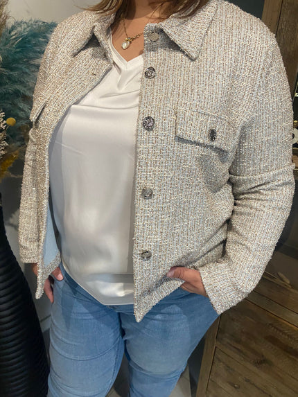 Iemand die de Amagia - Fabiola draagt, een ruimvallende blauw-beige blazer van elastische stof, over een witte satijnen blouse en een blauwe spijkerbroek, staat binnen met de handen in de zakken bij houten meubels en decoratieve planten.