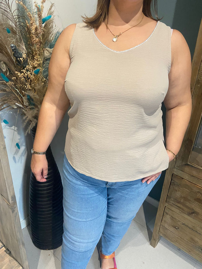 Een vrouw modelleert de An Vito - top in beige met een lichtblauwe jeans, binnen bij een grote vaas met gedroogde bloemen en pauwenveren. Ze straalt moeiteloos chic uit, haar linkerhand in haar zak en hoofd lichtjes gedraaid.