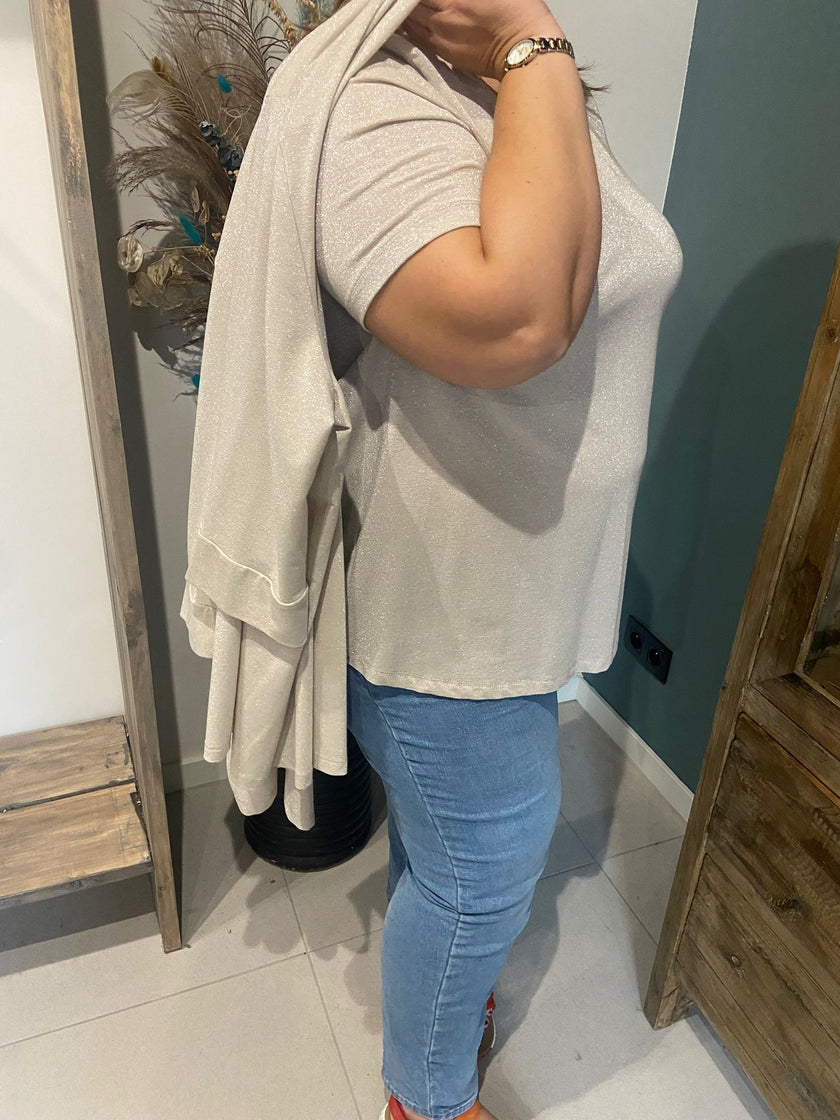 Een persoon in een lichte top met korte mouwen en een blauwe spijkerbroek staat opzij en houdt de Amagia - Fabiola blauw beige elastische blazer over zijn schouder. De blazer bedekt gedeeltelijk het gezicht en om de pols is een horloge zichtbaar.