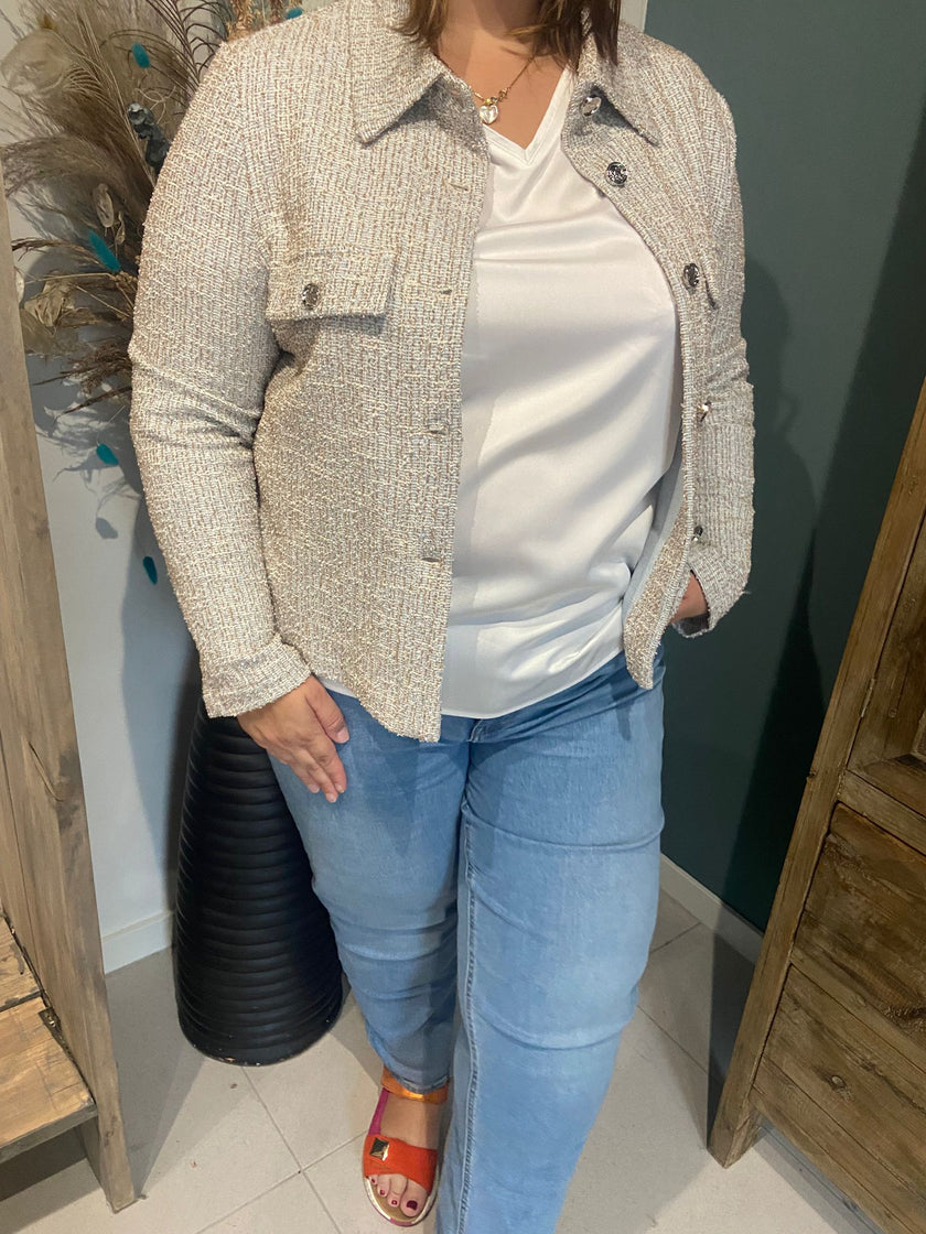 Een persoon die de Amagia - Fabiola blauw beige blazer van elastische stof draagt over een witte blouse, een lichtblauwe spijkerbroek en oranje sandalen staat binnen met één hand in zijn zak, omringd door gedroogde planten en houten meubels.