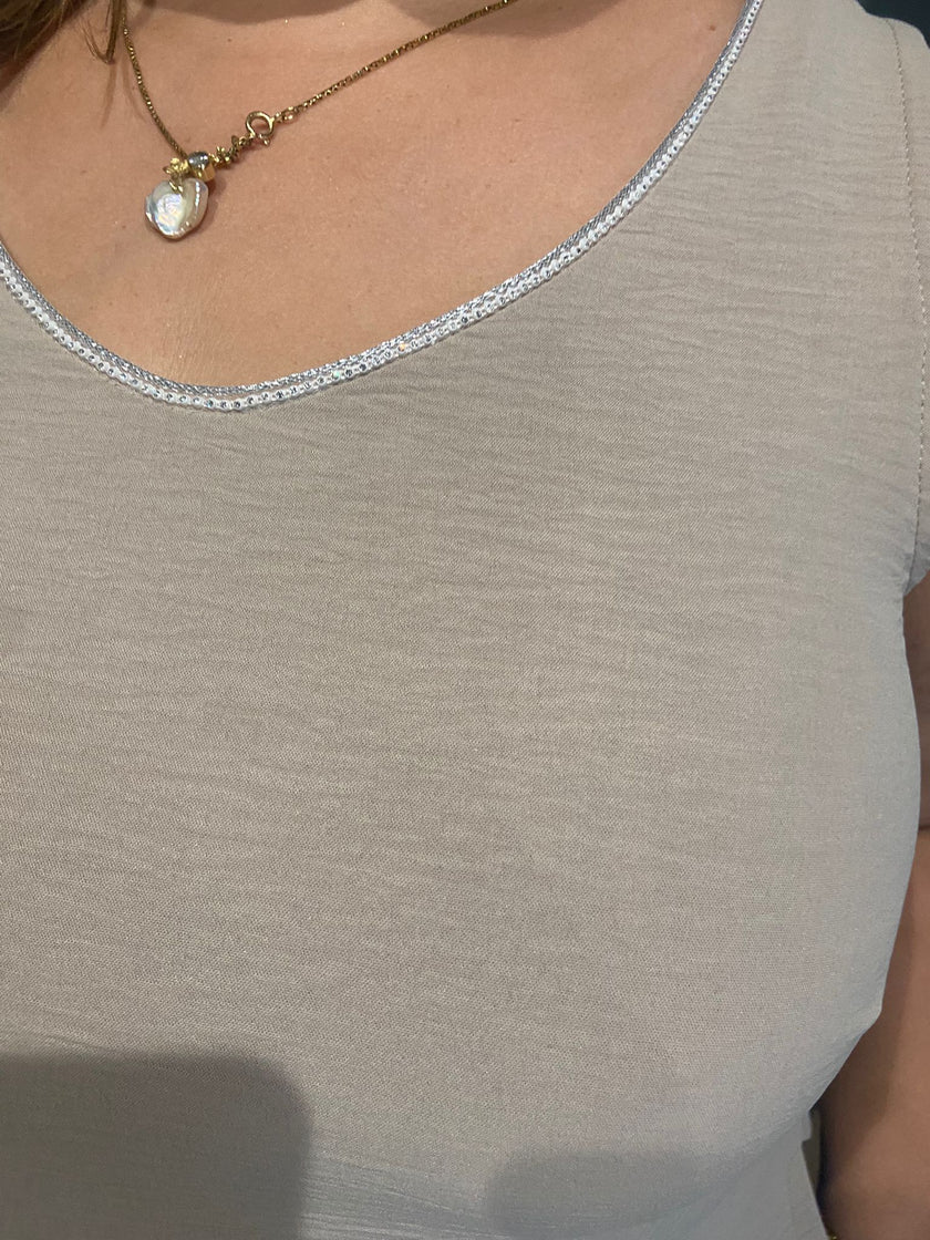 Een persoon draagt de An Vito - top in beige met een subtiel structuurpatroon en een gouden halsketting met een ronde hanger met een glanzende, iriserende steen, die de elegante glitterafwerking op de bovenborst en halslijn benadrukt.