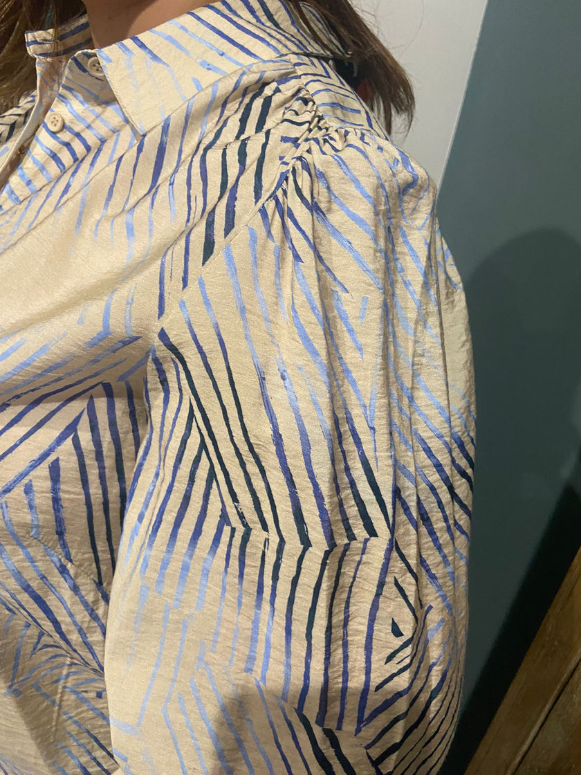 Een close-up van een persoon die de YEST Hemd draagt, een beige shirt met lange poff mouwen en onregelmatige blauwe en zwarte strepen, binnen bij een groen-witte muur.