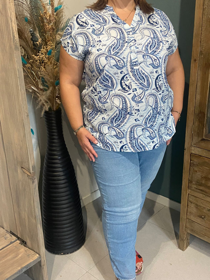 Een persoon staat binnen bij een hoge zwarte vaas met gedroogde bloemen, draagt het shirt rustique blauw en een lichtblauwe spijkerbroek. Hun hand zit in hun zak en hun gezicht is niet zichtbaar.