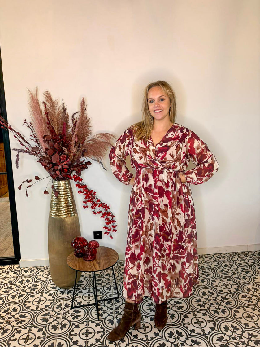 Een vrouw met het Kleedje - herfstblaadjes patroon, een ruime pasvorm jurk in rode en crèmekleurige tinten, staat binnen op een tegelvloer naast een hoge vaas met gedroogd gras en rood gebladerte en een kleine ronde tafel met rode glazen ornamenten.