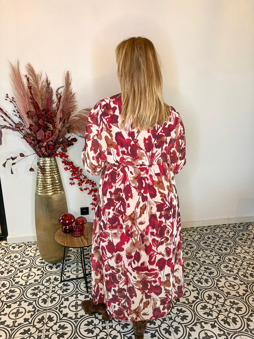 Een vrouw met blond haar staat met haar rug naar de camera en draagt het Kleedje - herfstblaadjes patroon in tinten rood, roze en wit. Ze staat binnen op een zwart-wit gedessineerde vloer naast een hoge vaas met gedroogde bloemen.