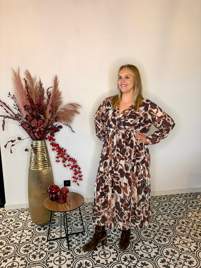 Een vrouw in een Kleedje - herfstblaadjes patroon staat glimlachend met een hand op haar heup naast een hoge vaas met gedroogde planten en een tafeltje met rode versiering, dit alles op een zwart-wit gedessineerde vloer.