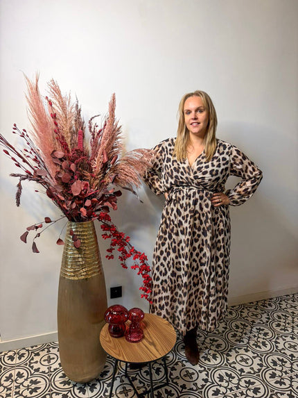 Een vrouw in een kleed - panterprint staat binnen naast een hoge vaas met gedroogde roze en beige bloemen, op vloertegels met patroon bij een kleine ronde tafel met rode glazen decoraties.