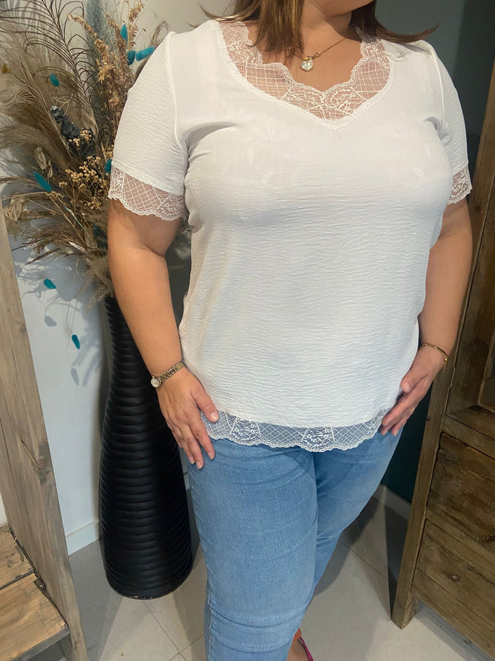 Een vrouw met het Ann Vito - shirt met kanten details en een lichtblauwe jeans staat binnen naast een hoge zwarte vaas gevuld met gedroogde bloemen en pauwenveren, met een hand op haar heup.