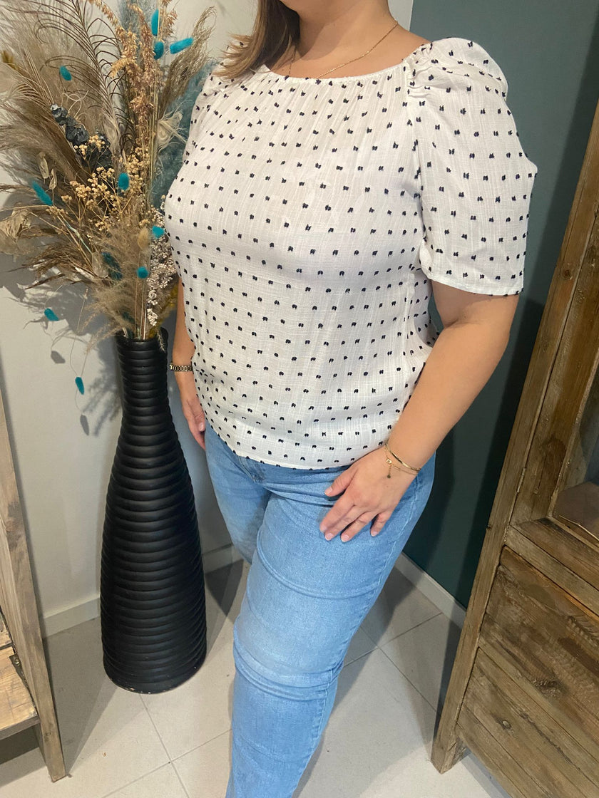 Een vrouw draagt het shirt - Korte mouwen - Wit/Stippen met een lichtblauwe spijkerbroek en staat binnen bij een hoge zwarte vaas met gedroogde planten. Haar gezicht is gedeeltelijk verborgen, wat de ontspannen damesmode benadrukt.