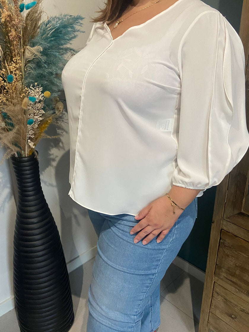 Een vrouw staat binnen naast een hoge zwarte vaas met gedroogde bloemen, draagt de Amagia - Grace off white blouse en een blauwe spijkerbroek. Haar gezicht is niet zichtbaar in het beeld.