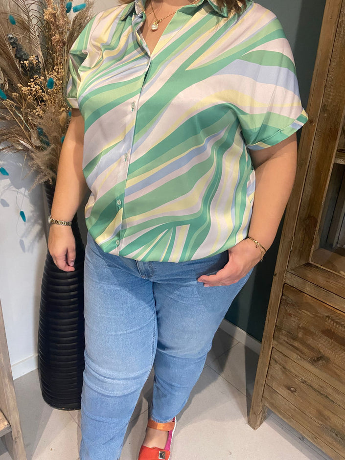 Een vrouw in een zomerse look draagt de Amagia - Hannah pastelgroen, geel en wit gestreept shirt met korte mouwen met lichtblauwe jeans, oranje schoenen en gouden sieraden, staand bij een houten kast en een hoge vaas met gedroogde bloemen.