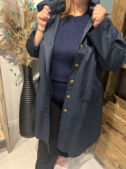 Binnen draagt een persoon de Trenchcoat - Zwart - Blauw - Bruin over een fonkelend blauw topje en een zwarte broek terwijl hij de kraag vasthoudt; een grote zwarte vaas met gedroogde bloemen verschijnt op de achtergrond.