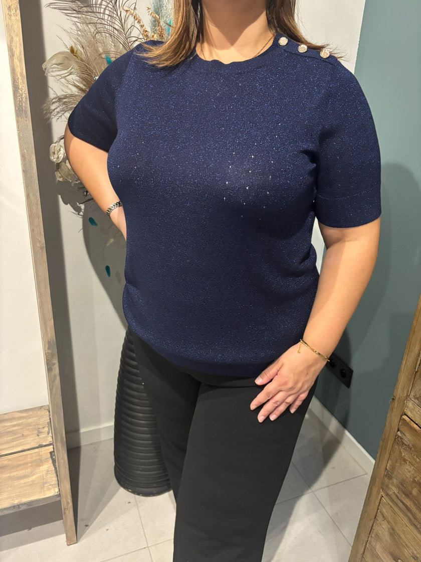 Een vrouw binnenshuis draagt het Floortje - Donker marine blousje met glinsterende schouderknopen en een zwarte broek. Eén hand rust op haar heup terwijl ze gedeeltelijk naar de camera kijkt.