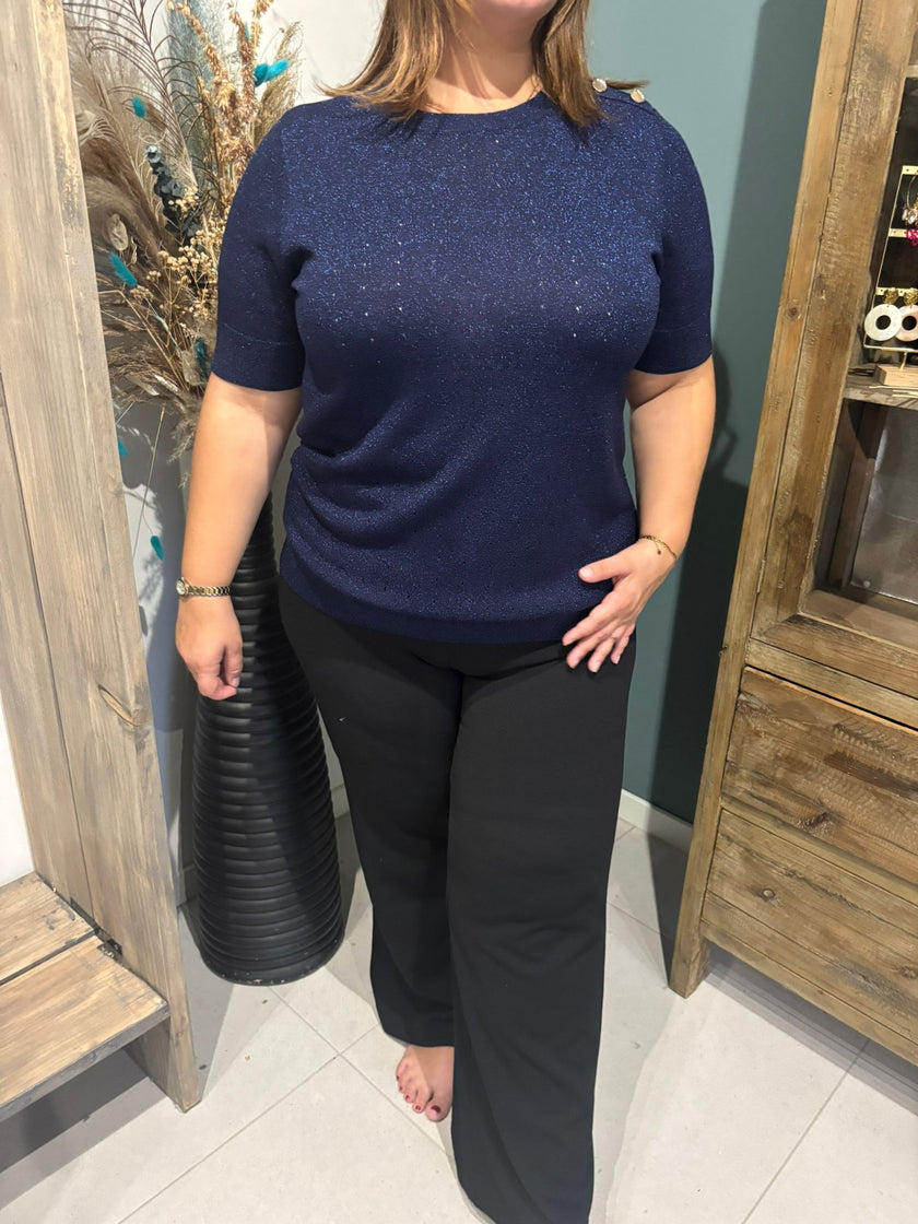 Een vrouw staat blootsvoets binnen naast houten meubels, ze draagt het Floortje - Dark Navy glitterblousje met korte mouwen en een zwarte broek. Haar gezicht is gedeeltelijk buiten beeld en een decoratieve vaas met gedroogde planten staat vlakbij.