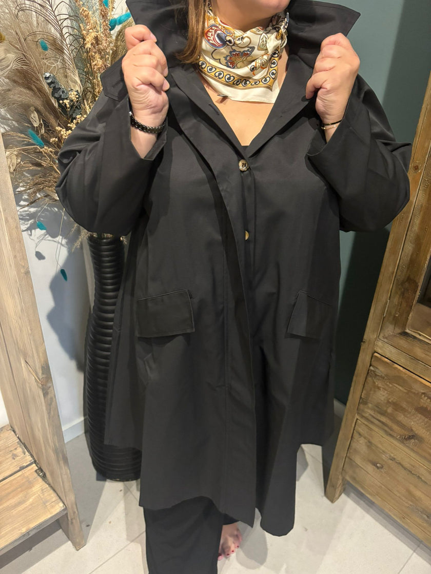 Een persoon met de Trenchcoat - Zwart - Blauw - Bruin en een zijden sjaal met patroon staat binnen en houdt de kraag van de jas omhoog. Alleen het ondergezicht en de handen zijn zichtbaar, met houten meubels en decoratieve planten op de achtergrond.