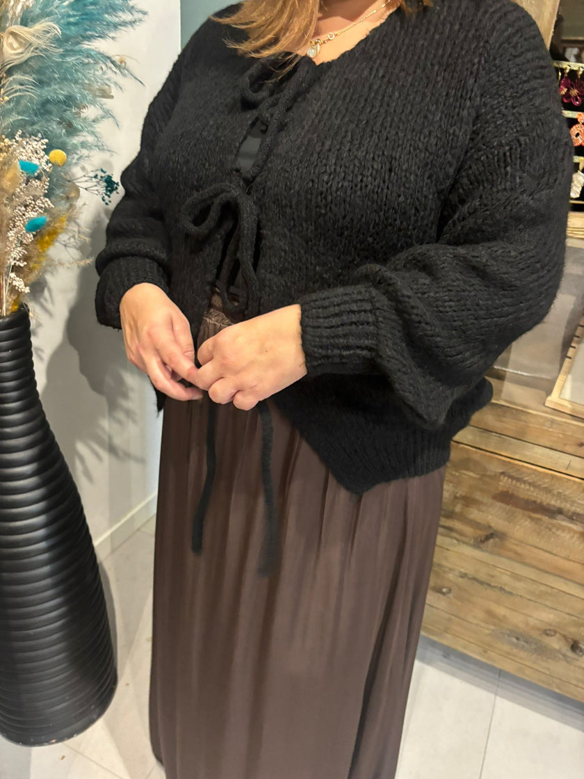 Een persoon met de Cardigan - Zwart - Bordeaux, een grof gebreide zwarte trui met opvallende knoopdetails en een lange bruine rok staat binnen bij een zwarte vaas met gedroogde bloemen en toont een vrouwelijke uitstraling.