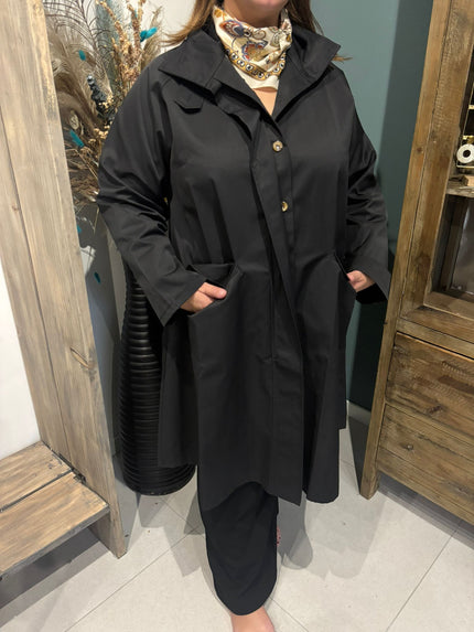 Een persoon in een Trenchcoat - Zwart - Blauw - Bruin met gouden knopen en een sjaal met beige patroon staat binnenshuis, handen in zakken, bij houten meubels en gedroogde planten.