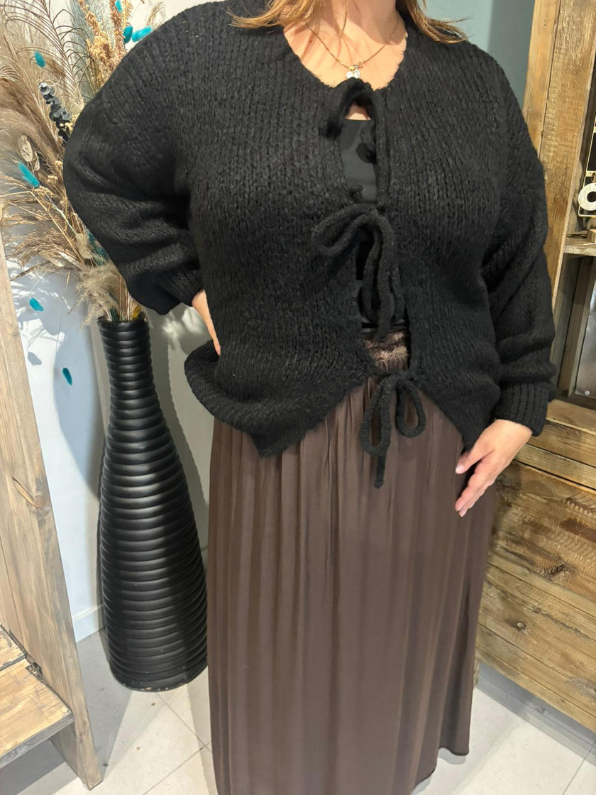 Een persoon draagt de Cardigan - Zwart - Bordeaux over een bruine plooirok en staat binnenshuis naast een hoge zwarte vaas met decoratieve gedroogde grassen voor een vrouwelijke uitstraling.