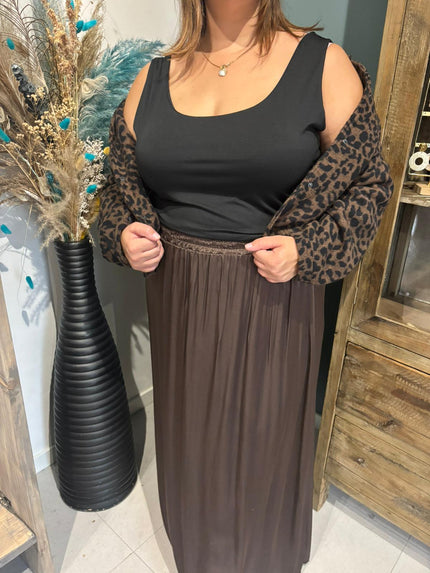 Een vrouw draagt een Zwart topje, een bruin vest met luipaardprint over haar schouders en een lange bruine rok (TU een maat 48-54). Ze staat binnen naast een hoge zwarte vaas met gedroogde bloemen en een houten kast.