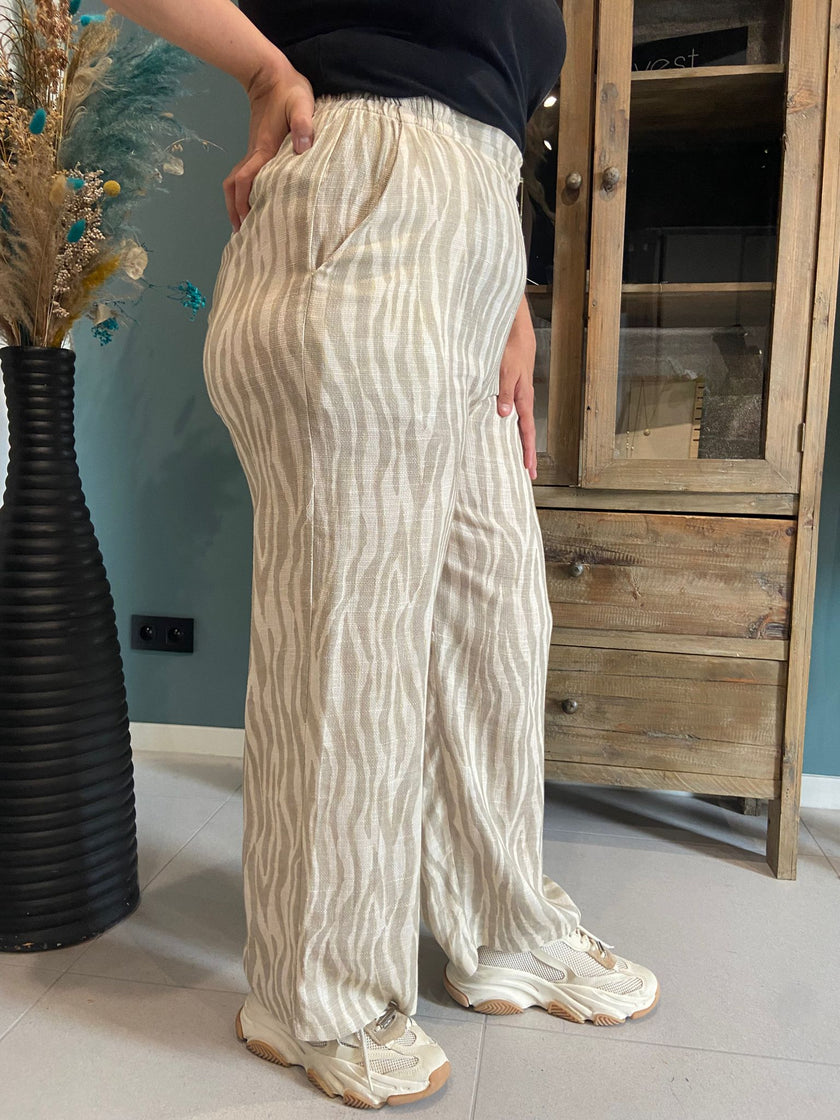 Een persoon binnenshuis draagt de An Vito - Beige/wit gestreepte broek met wijde pijpen en crème sneakers, hand in zak, staand naast een zwarte vaas met gedroogde bloemen en een houten kast.