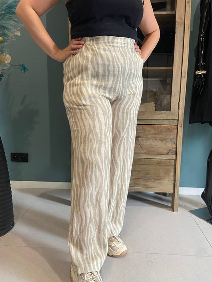 Een persoon die de An Vito - Beige/wit gestreepte, luchtige broek met een subtiel golvend streeppatroon, een zwarte mouwloze top en witte sneakers draagt, staat binnen met de handen op de heupen.