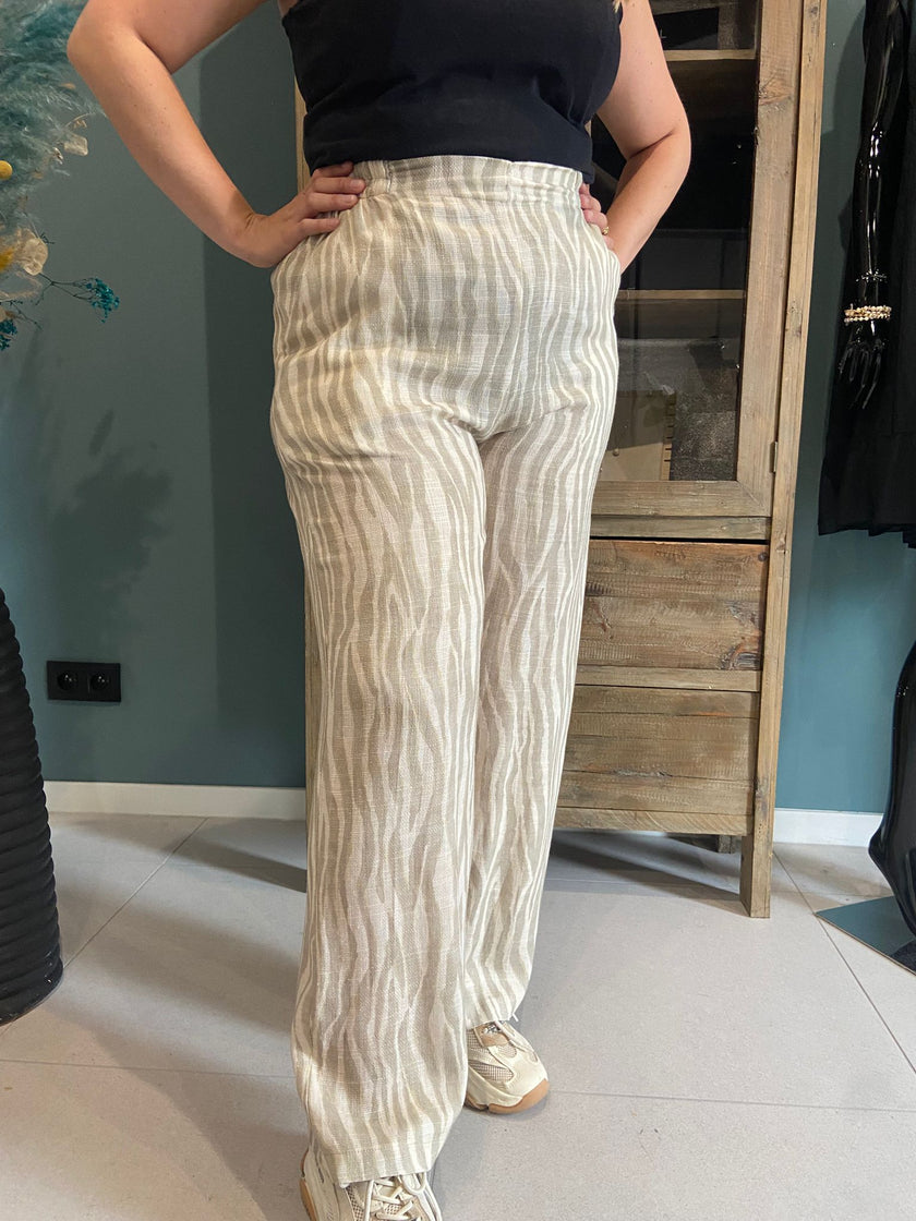 Een persoon die de An Vito - Beige/wit gestreepte, luchtige broek met een subtiel golvend streeppatroon, een zwarte mouwloze top en witte sneakers draagt, staat binnen met de handen op de heupen.