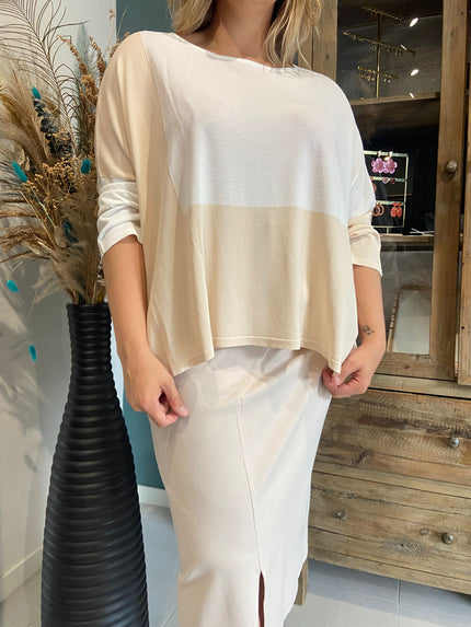 Een vrouw met de Amagia - Emilie damesblouse - een losse crèmekleurige top met lange mouwen - en een bijpassende rok met split aan de voorkant staat binnen naast een hoge zwarte vaas met gedroogde planten en pauwenveren.