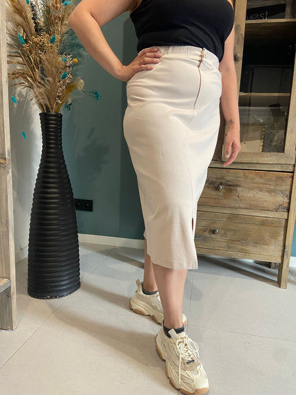 Een vrouw draagt de Amagia - Gemma high-waisted beige kokerrok met split, een zwart topje en grove beige sneakers bij een houten kast en een hoge zwarte vaas met gedroogde bloemen.