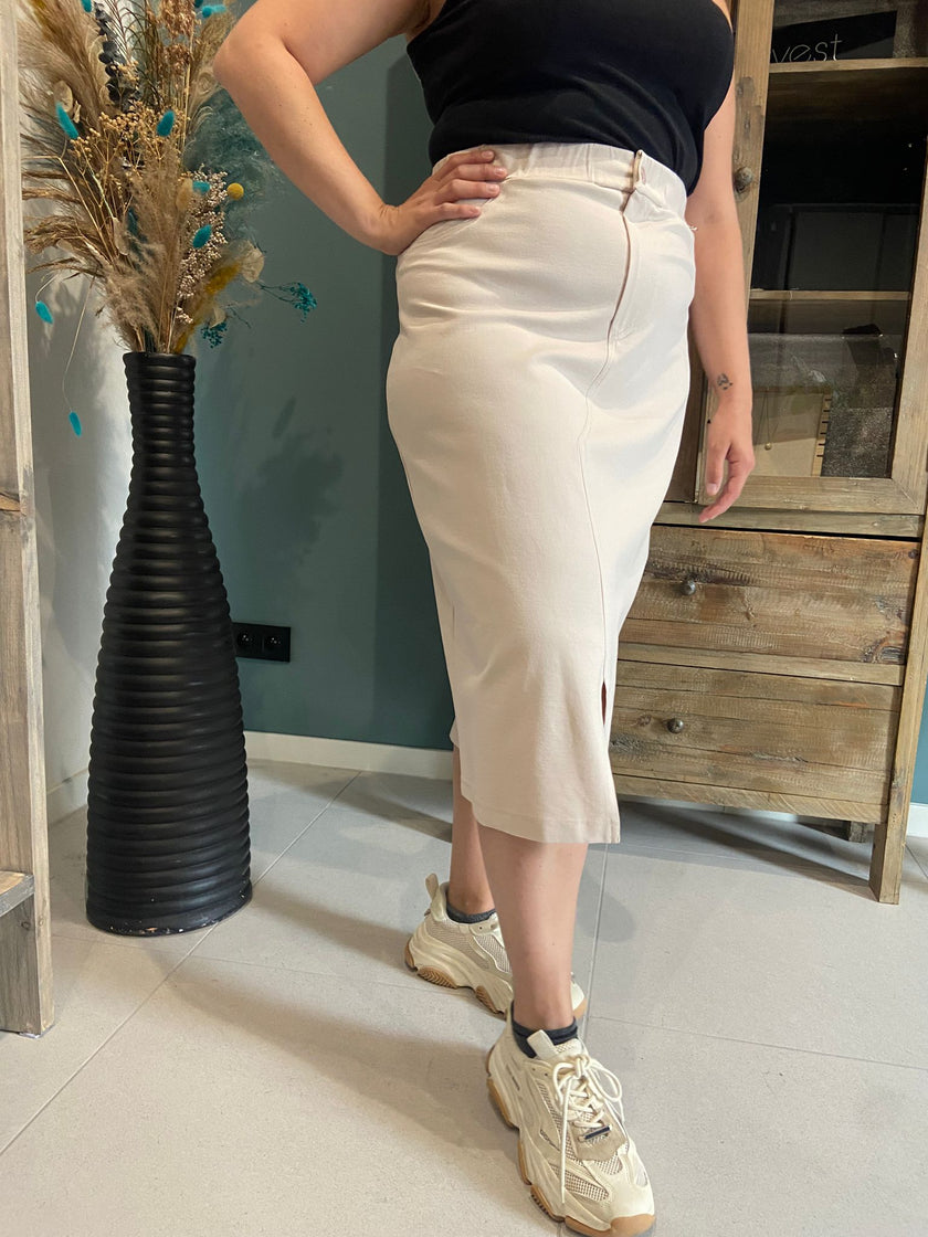 Een vrouw draagt de Amagia - Gemma high-waisted beige kokerrok met split, een zwart topje en grove beige sneakers bij een houten kast en een hoge zwarte vaas met gedroogde bloemen.