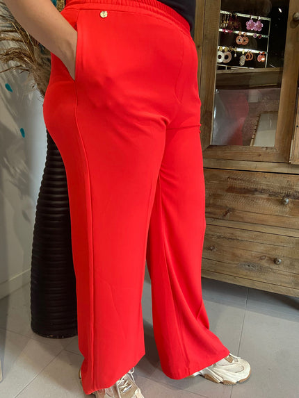 De persoon draagt de koraalrode Italiaanse broek van Amagia - Grosseto met crèmekleurige sneakers en de handen in de zakken. Hij staat binnen bij een houten kast, waarbij alleen de onderste helft zichtbaar is.