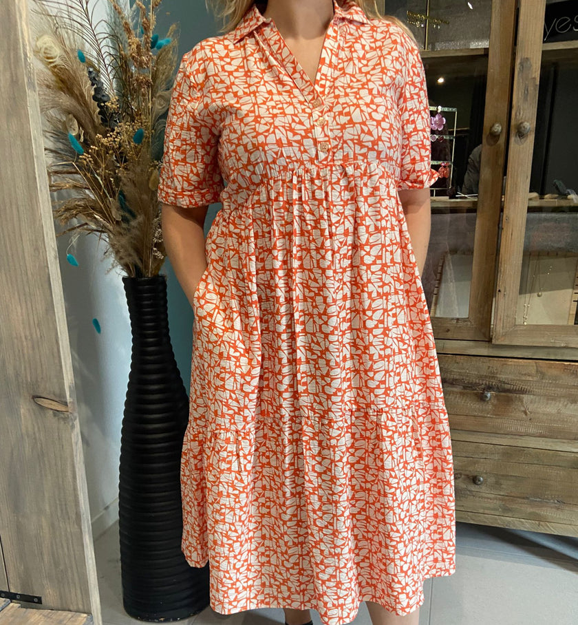 Een vrouw staat binnen en draagt Kleedje - Oranje, een oranje met wit gedessineerde zomerjurk met zakken. Ze staat bij een zwarte vaas met gedroogde planten, met houten meubels op de achtergrond. Haar gezicht is gedeeltelijk buiten beeld.