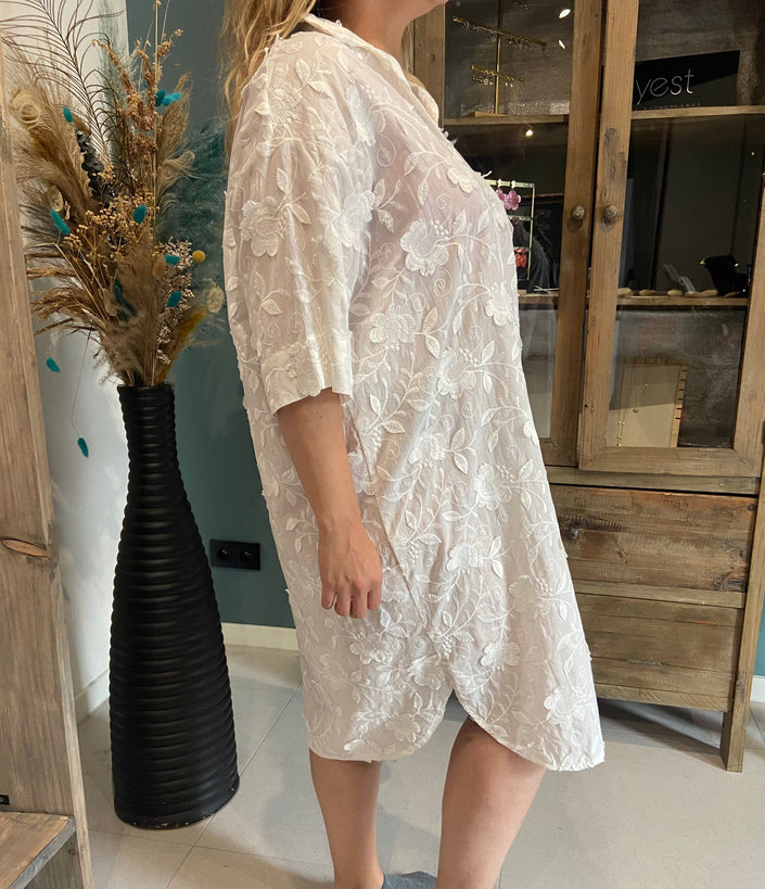 Een persoon in de Amagia - Gisele zomers wit kleedje (maat 44, kanten afwerking) staat binnen bij een houten kast en een hoge zwarte vaas met gedroogde bloemen, van opzij gezien.