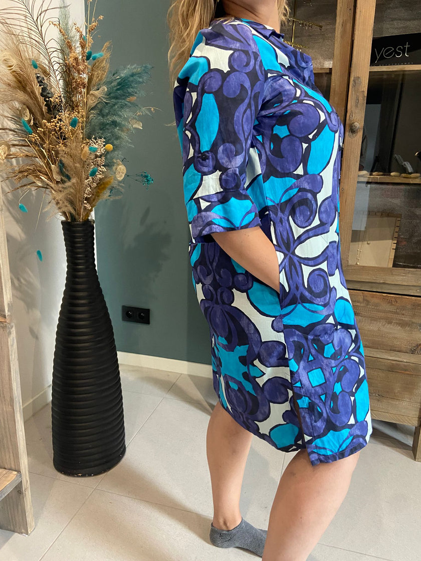 Binnen kijkt een persoon opzij in de An Vito - 4750: een knielange blauwe jurk met blauwe en paarse patronen en opgerolde mouwen, een hand in een zak. Een hoge vaas met gedroogde bloemen staat op de achtergrond.