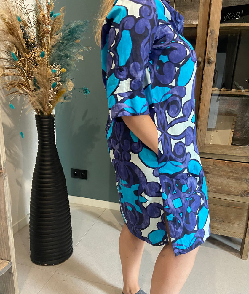 Een persoon staat binnen en draagt de An Vito - 4750 blauwe jurk met een levendig blauw en paars patroon, handen in de zakken. Naast hen staat een hoge zwarte vaas met gedroogde bloemen bij een houten kast tegen een groenblauwe muur.