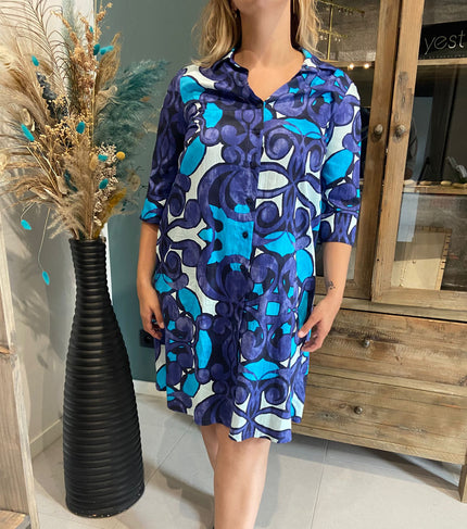 Een vrouw in een blauwe jurk met patroon staat binnen bij een hoge zwarte vaas met gedroogde bloemen en een rustieke houten kast, met An Vito - 4750.