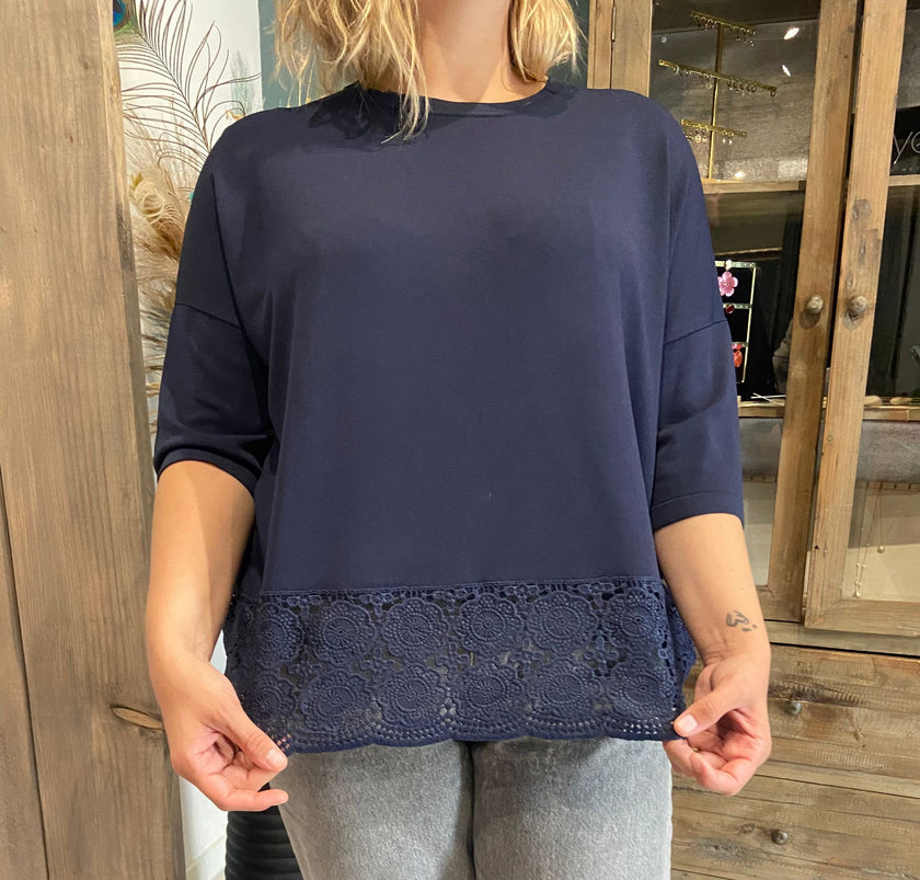 Een persoon die de Blouse - Blauw draagt, een loszittende top met kanten details, in combinatie met een lichtgrijze broek, staat binnen voor een houten kast.