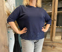 Een vrouw staat binnen, handen op de heupen, draagt de Blouse - Blauw met kanten zoom en een lichtgrijze jeans. Op de achtergrond staan houten meubels en een hoge vaas gevuld met pauwenveren.