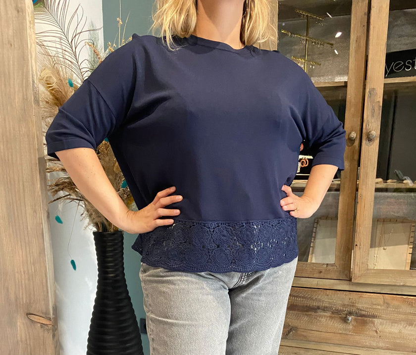 Een vrouw staat binnen, handen op de heupen, draagt de Blouse - Blauw met kanten zoom en een lichtgrijze jeans. Op de achtergrond staan houten meubels en een hoge vaas gevuld met pauwenveren.