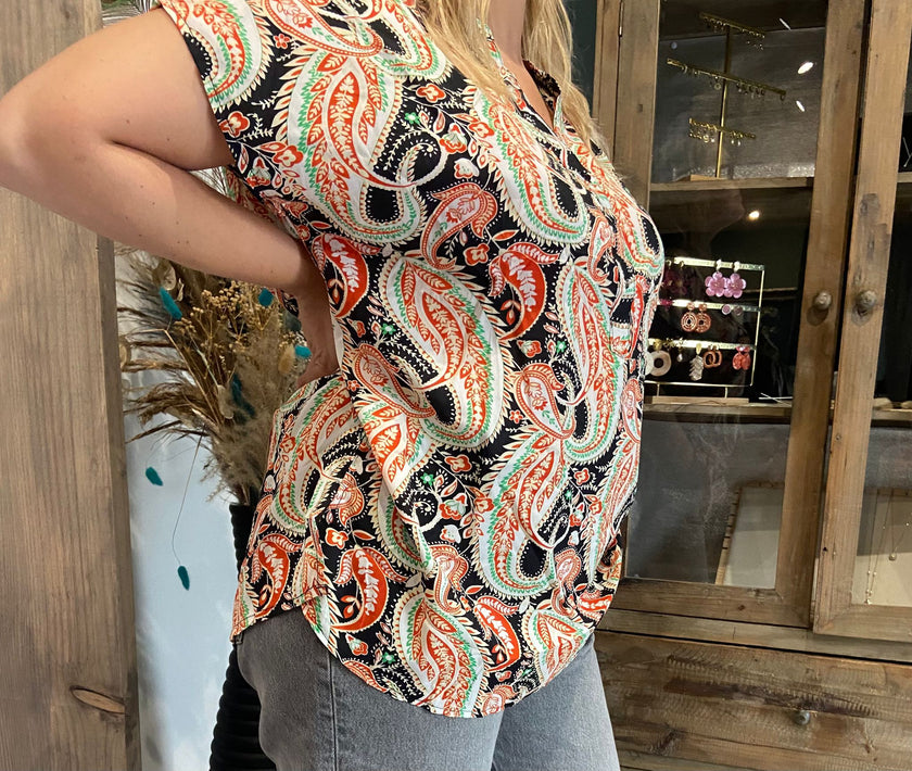 Een persoon in een T-shirt met kleurrijke print en een grijze spijkerbroek staat binnen bij een houten kast met sieraden en decor, wat een charmante boho-look creëert.
