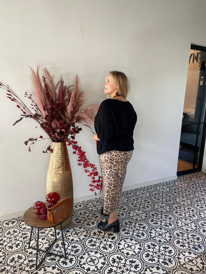 Een vrouw staat binnen op een tegelvloer met patroon, met haar gezicht weg van de camera. Ze draagt een zwarte trui en de Zweedse luipaardrok. Naast haar staat een hoge gouden vaas met bordeauxrode gedroogde planten en een tafeltje met rood decor.