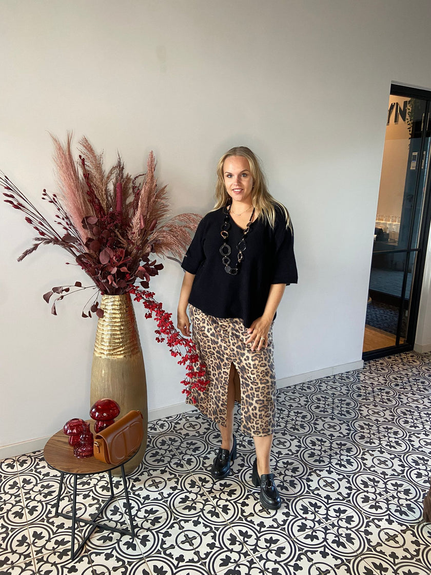 Een vrouw staat binnen op tegels met een patroon en draagt een zwart topje, een Zweedse luipaardrok en zwarte schoenen. Ze poseert bij een hoge vaas met gedroogde bordeauxrode planten en naast een tafeltje met een bruine tas en rode ornamenten.