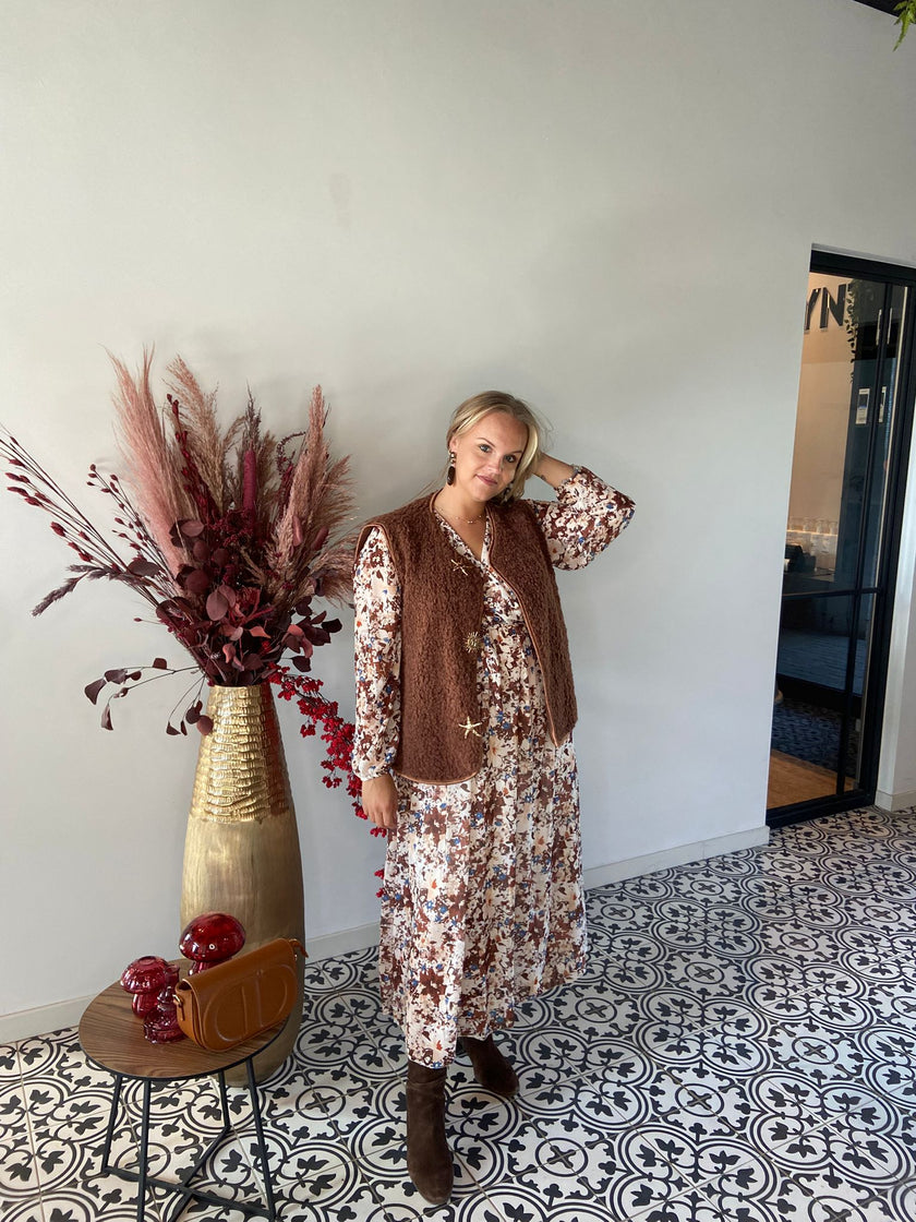 Een vrouw in een lange bloemetjesjurk, bruine laarzen en het Gilet met ster-en zon staat binnen op tegels met een patroon naast een hoge vaas met gedroogde bladeren en een tafeltje met een bruine tas en decoratieve voorwerpen.