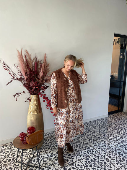 Een vrouw in een Gilet met ster-en zon over een gebloemde jurk staat binnen op tegels met sterren- en zonnenpatroon, terwijl ze haar haar aanraakt. Naast haar staat een gouden vaas met gedroogde bordeauxrode planten en een tafel met een rode handtas-herfst/winter 2025/2026.