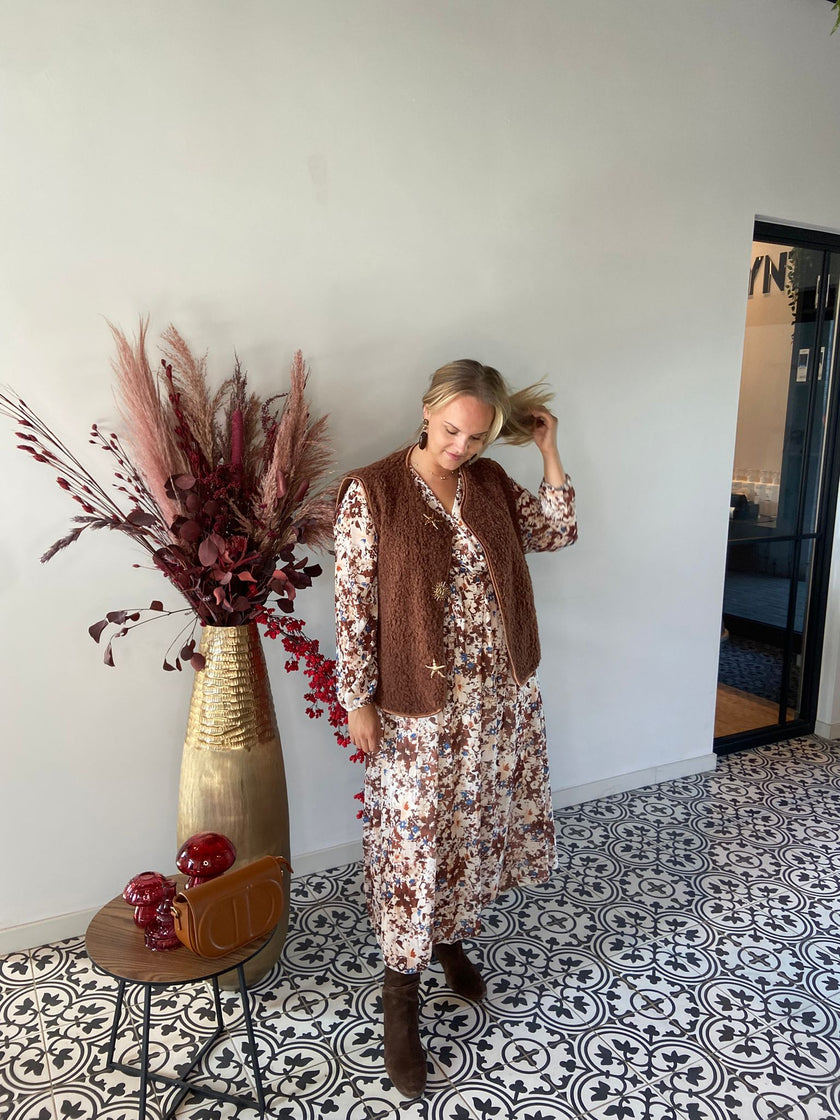 Een vrouw in een Gilet met ster-en zon over een gebloemde jurk staat binnen op tegels met sterren- en zonnenpatroon, terwijl ze haar haar aanraakt. Naast haar staat een gouden vaas met gedroogde bordeauxrode planten en een tafel met een rode handtas-herfst/winter 2025/2026.