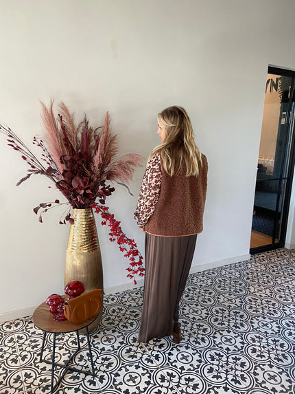 Een vrouw met golvend blond haar kijkt naar een effen muur, draagt een Bruine rok met een elastische tailleband en een bruin en beige jasje. Naast haar staat een hoge gouden vaas met gedroogde rode planten en een tafeltje met decoratieve voorwerpen.
