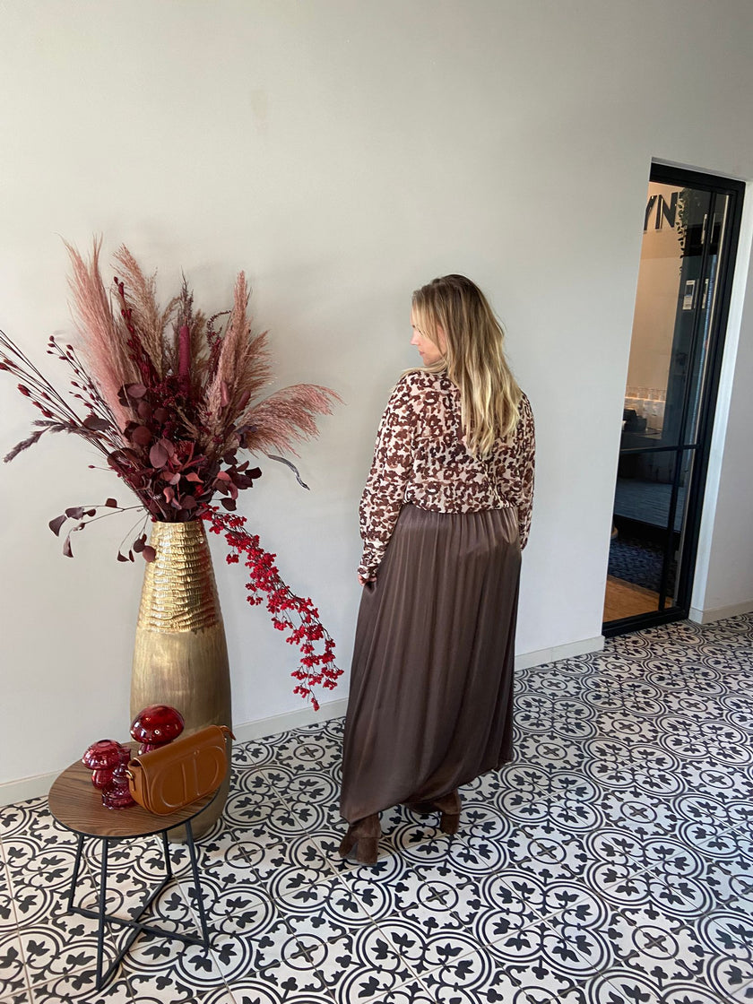 Een vrouw met lang blond haar in een shirt met print - beschikbaar in 2 kleuren en een lange bruine rok staat bij een hoge gouden vaas met rode en bruine gedroogde planten op zwart-witte tegels met patroon, die tijdloze elegantie uitstralen.