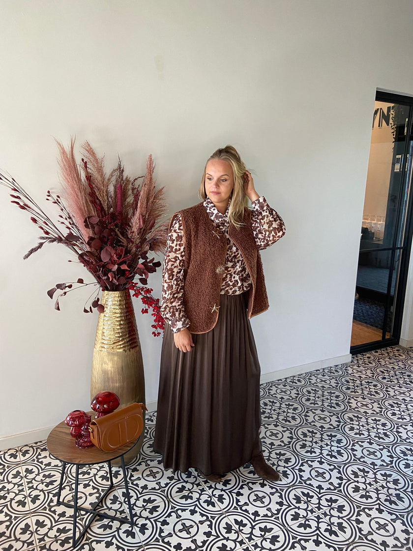 Een vrouw in een bruin vest, blouse van viscose met patroon en een Bruine rok met elastische tailleband staat binnen op zwart-witte tegels, naast een hoge vaas met gedroogde bloemen en een tafeltje met een handtas en rode appels.