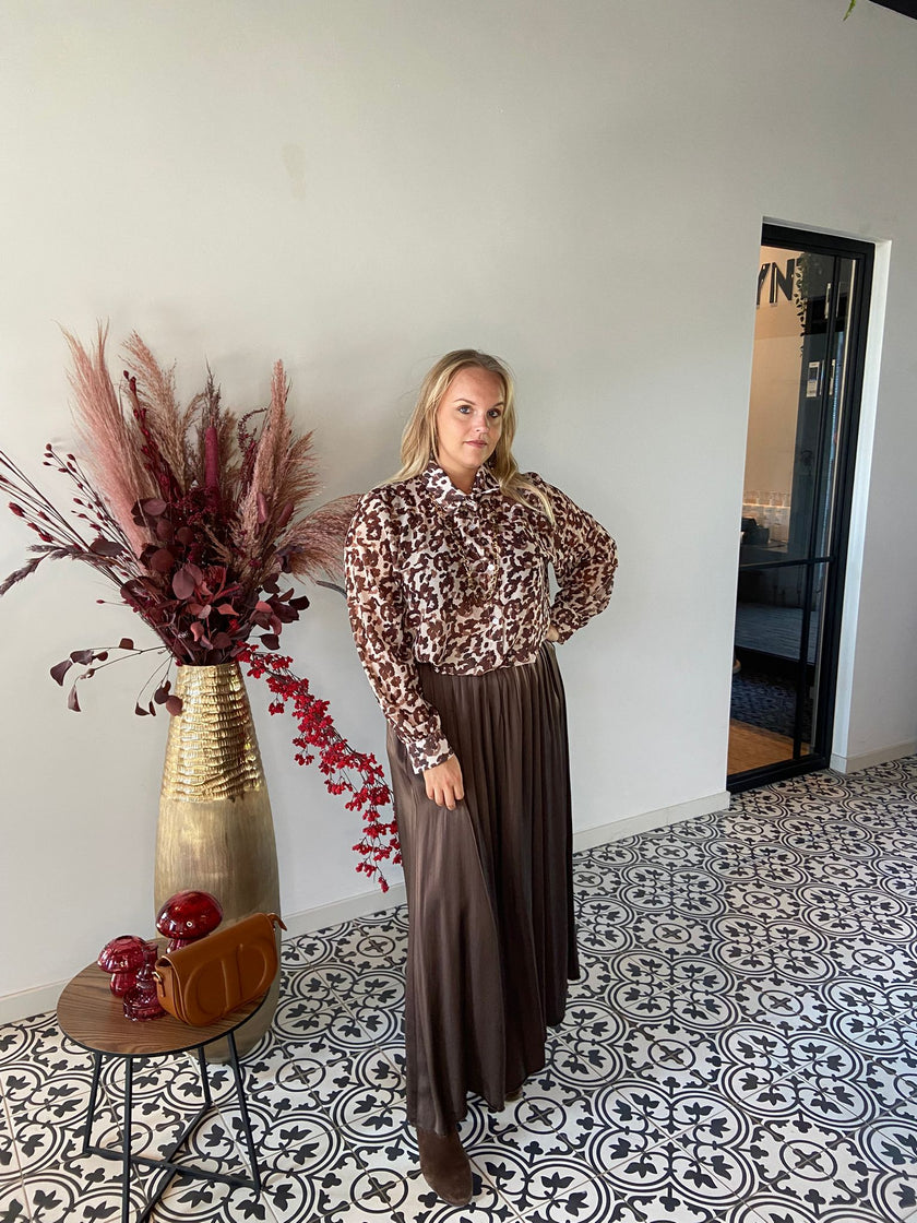 Een vrouw staat binnen op een tegelvloer met patroon en straalt tijdloze elegantie uit in het shirt met print - beschikbaar in 2 kleuren en een lange bruine rok. Ze poseert bij een ronde tafel met een bruine tas en gedroogd rood en roze gebladerte.